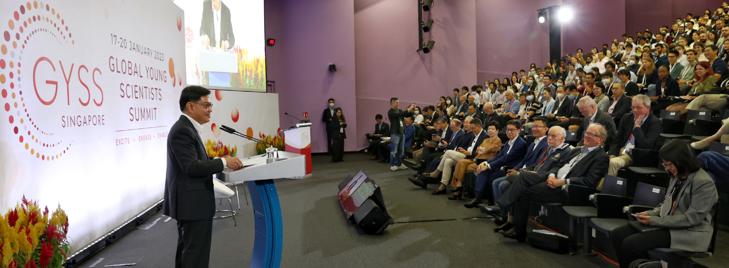 Man at podium speaking to a large audience, "GYSS Global Young Scientists Summit" backdrop.
